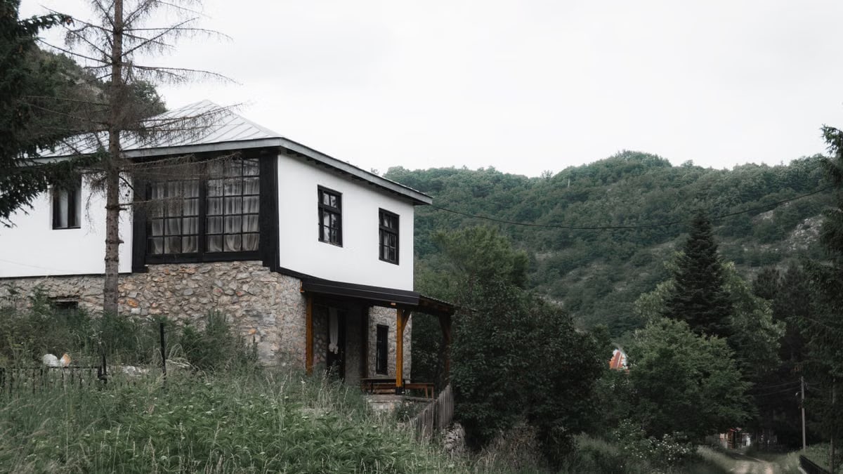 A charming house in a rural setting with lush green hills, trees, and a dirt road, perfect for peaceful retreats.